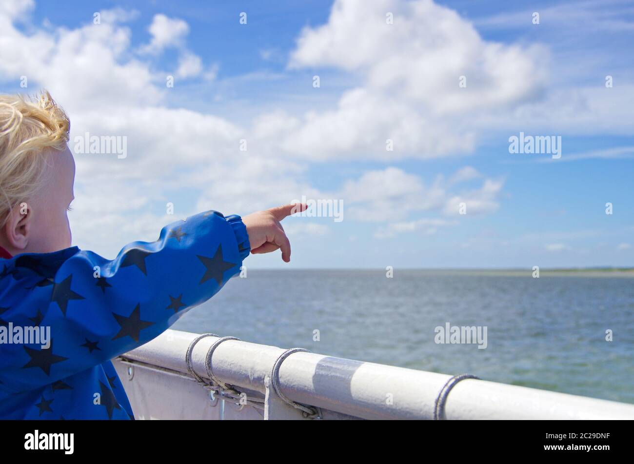 Ein Kleinkind auf einer Fähre zeigt mit einem Finger in Richtung Insel und blauem Himmel mit Wolken Stock Photo