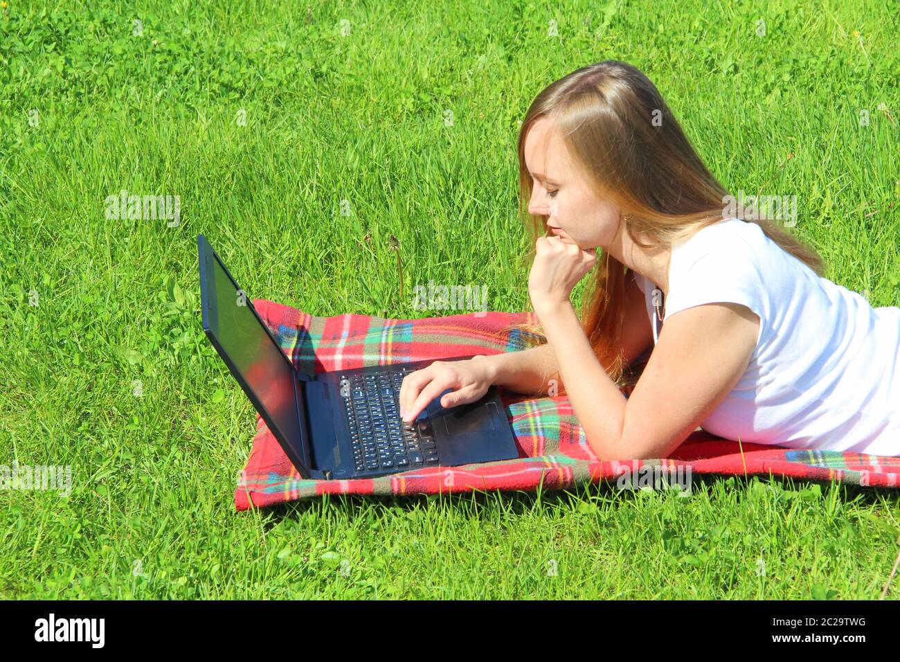 A beautiful young white girl in a white T-shirt and with long hair lies on a red plaid, on green grass, on the lawn and working behind a black laptop. Stock Photo