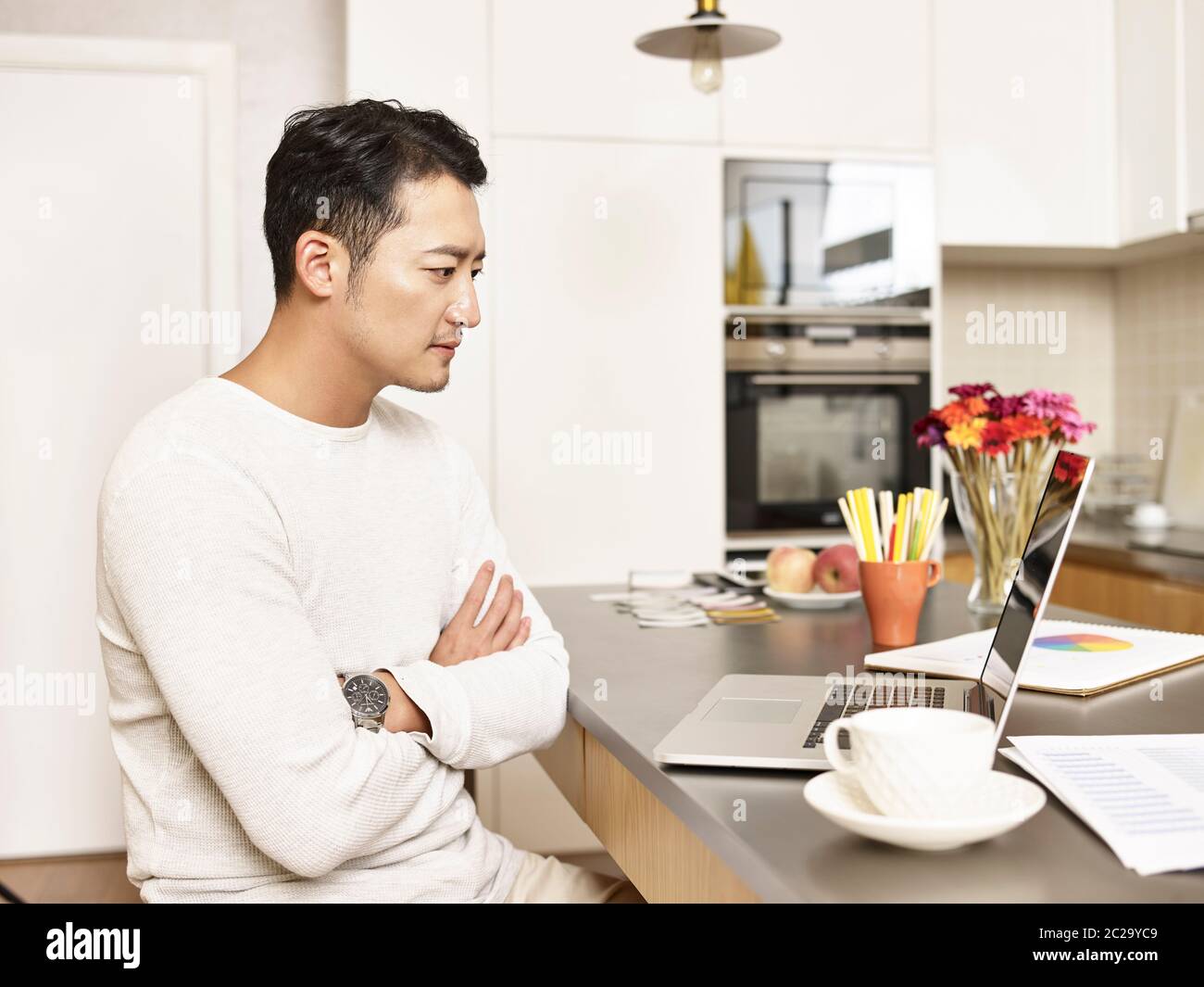 young asian businessman working from home sitting at kitchen counter looking at laptop computer arms crossed Stock Photo