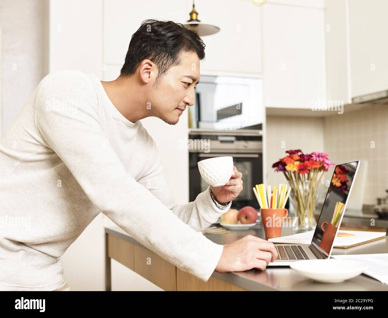 young asian businessman working from home standing by kitchen counter holding a cup of coffee looking at laptop computer Stock Photo