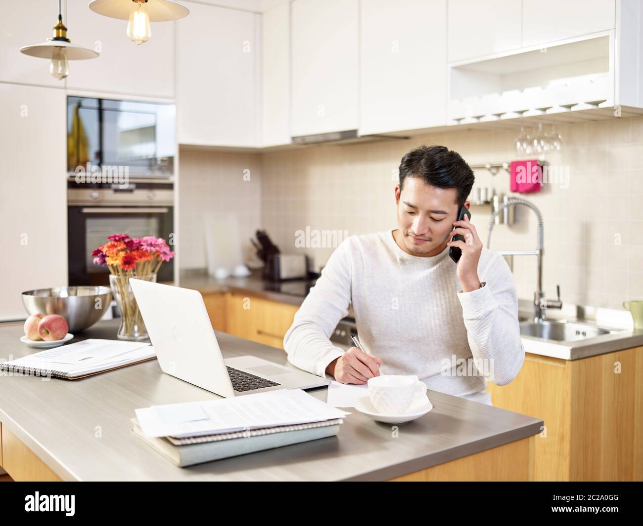 young asian business man sitting kitchen counter working at home talking on cellphone Stock Photo