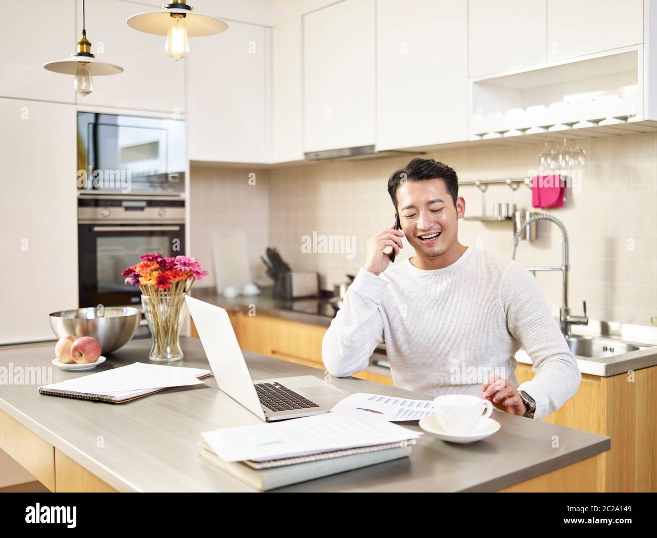 young asian business man sitting kitchen counter working at home talking on mobile phone laughing Stock Photo