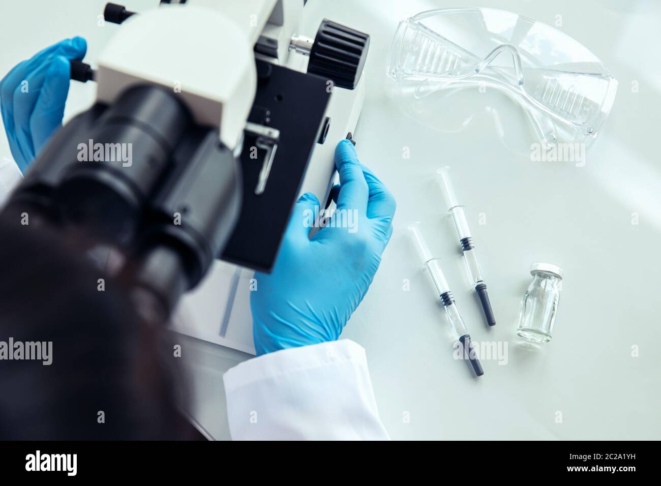 Female scientist uses microscope. Female scientist in protective gloves uses medical microscope with syringes, protective glasses and phial next to it Stock Photo