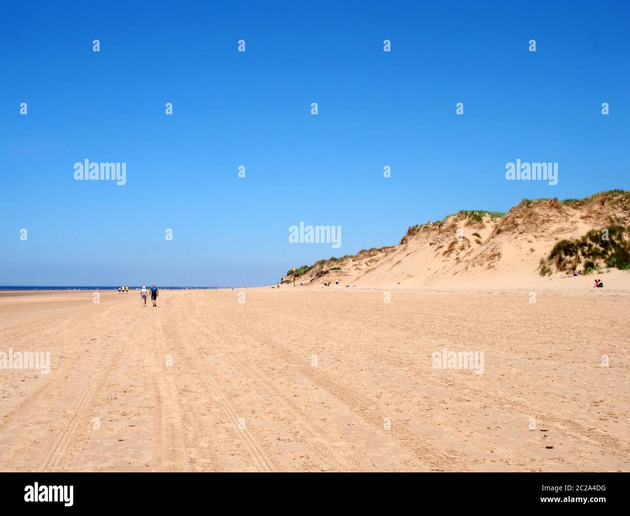 people walking on the beach with grass on the top of tall sand dunes on the sefton coast in merseyside with blue summer sea and Stock Photo