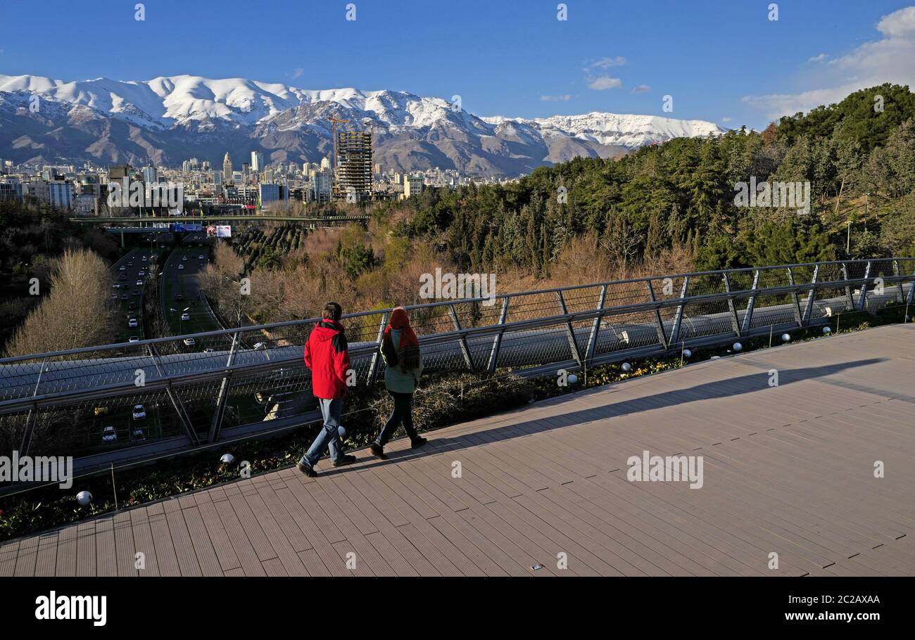 Iranian people crossing the modern Tabiat pedestrian bridge, to Taleghani Park, with the Alborz snow mountains in the background, in Teheran. Stock Photo