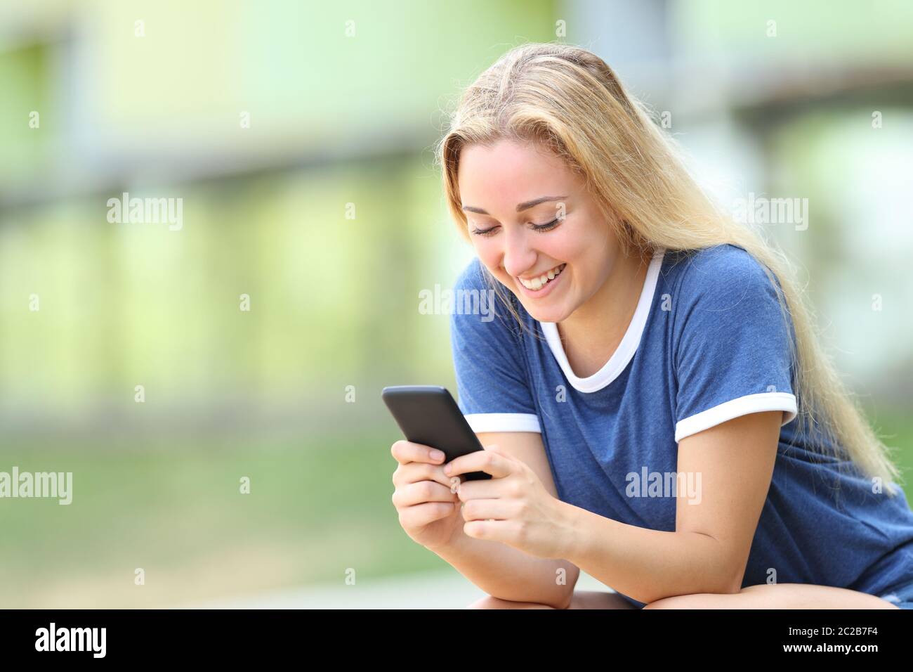 Happy teenage girl texing messages on smart phone sitting in an university campus Stock Photo