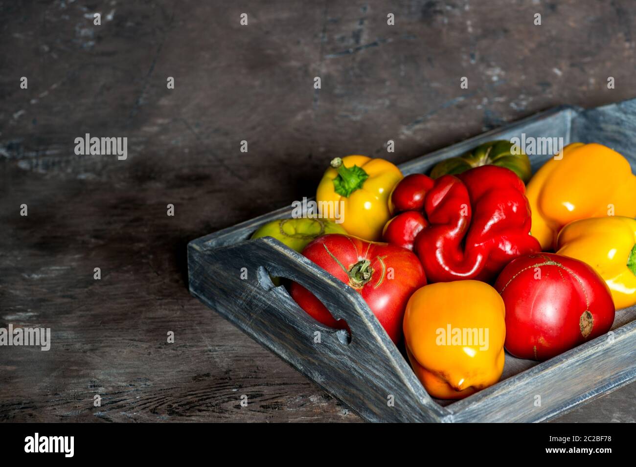 Imperfect natural peppers and tomatoes on an old wooden tray on a dark background. Copy Space. Stock Photo