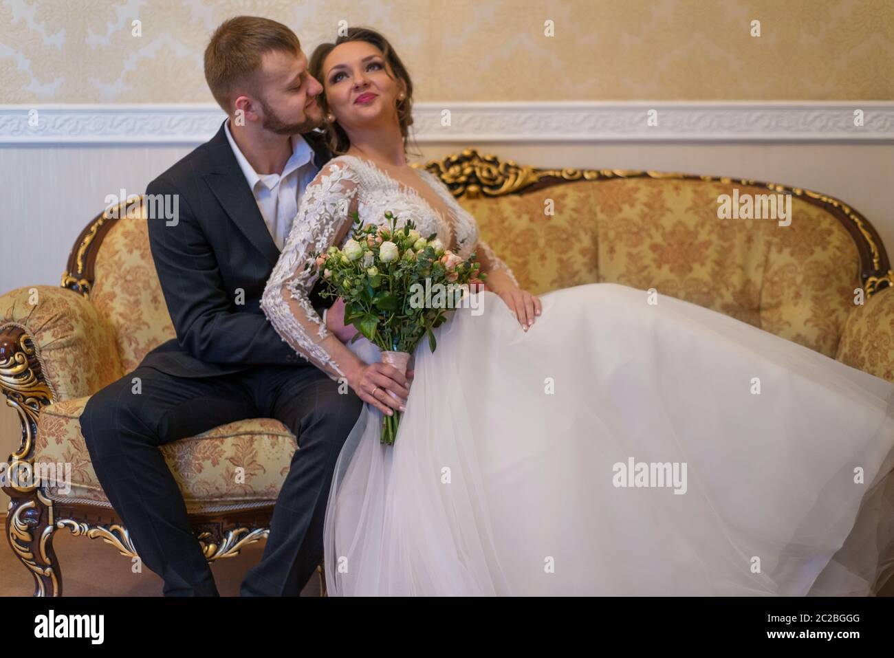 The bride and groom are sitting on a luxurious sofa, spreading a dress, the bride looks up, the bearded groom kisses her. Smiling young people of twen Stock Photo