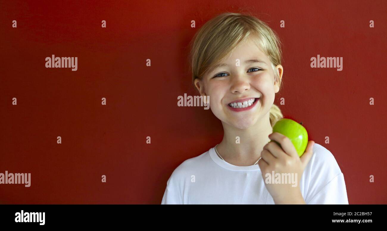 Happy blond little girl with green apple by the red wall Stock Photo