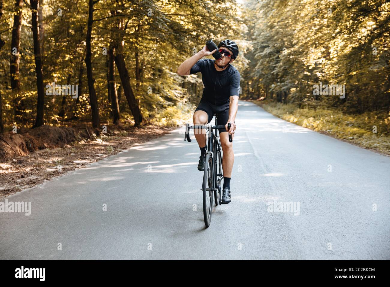 Professional sportsman in active wear and protective helmet drinking water while riding bike on fresh air. Mature man in mirrored glasses enjoying fav Stock Photo