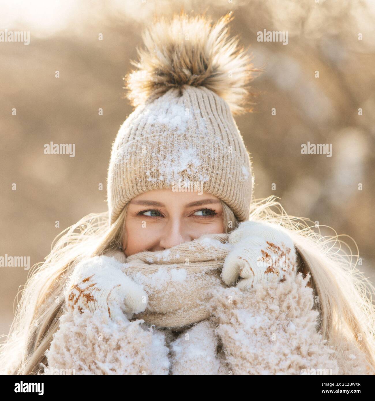 Portrait of pretty young woman with long hair in beige hat with fur pompon, warm scarf, coat, white gloves covered with snow at sunny winter day. Fema Stock Photo
