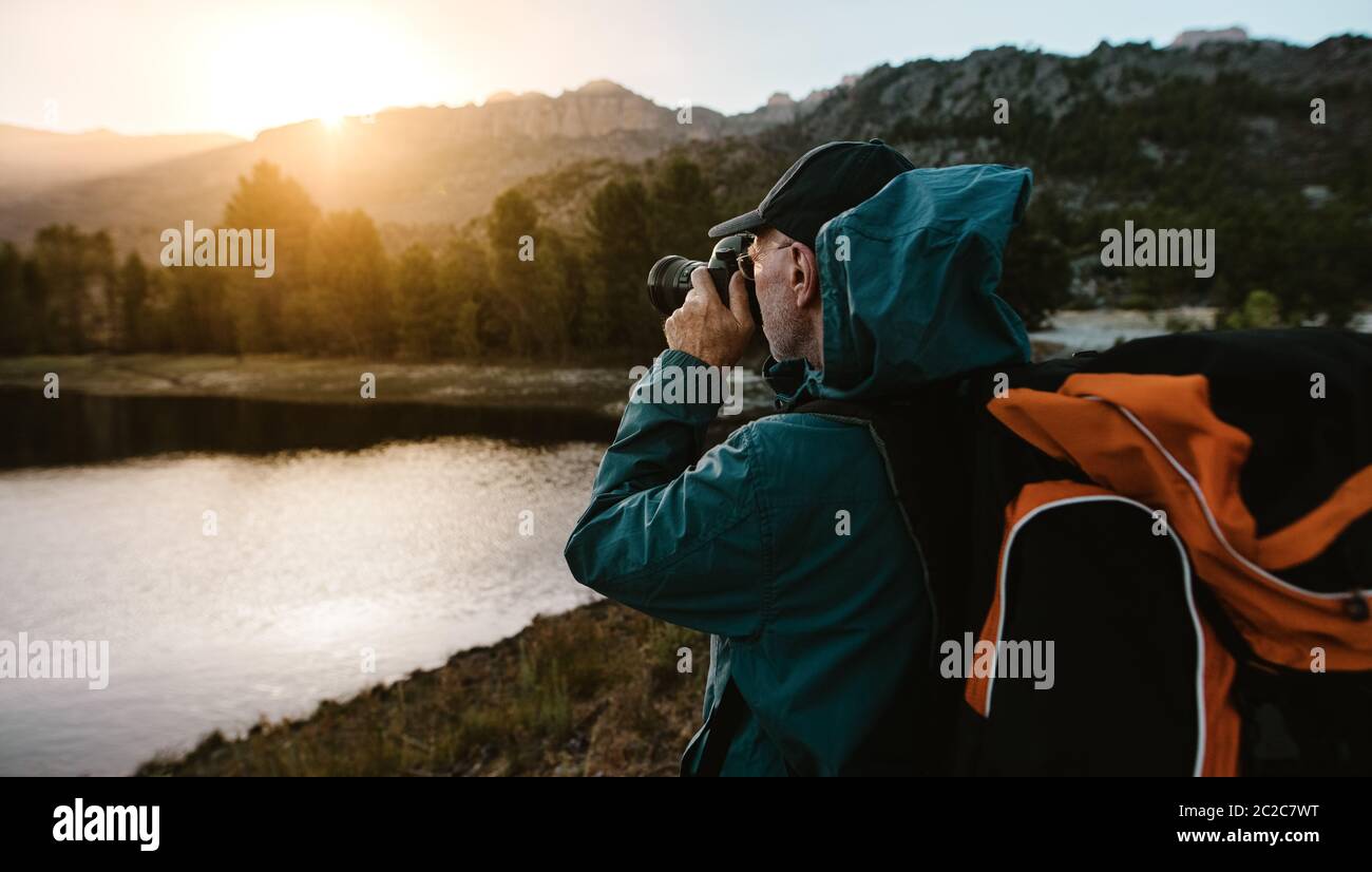 Senior man on hiking trip taking photographs of the view with a digital camera. Man hiker standing by the river in forest and taking photos. Stock Photo