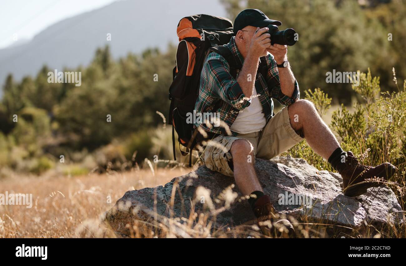 Senior man photographing with a digital camera. Retired man sitting on a rock and taking pictures on hiking trip. Stock Photo
