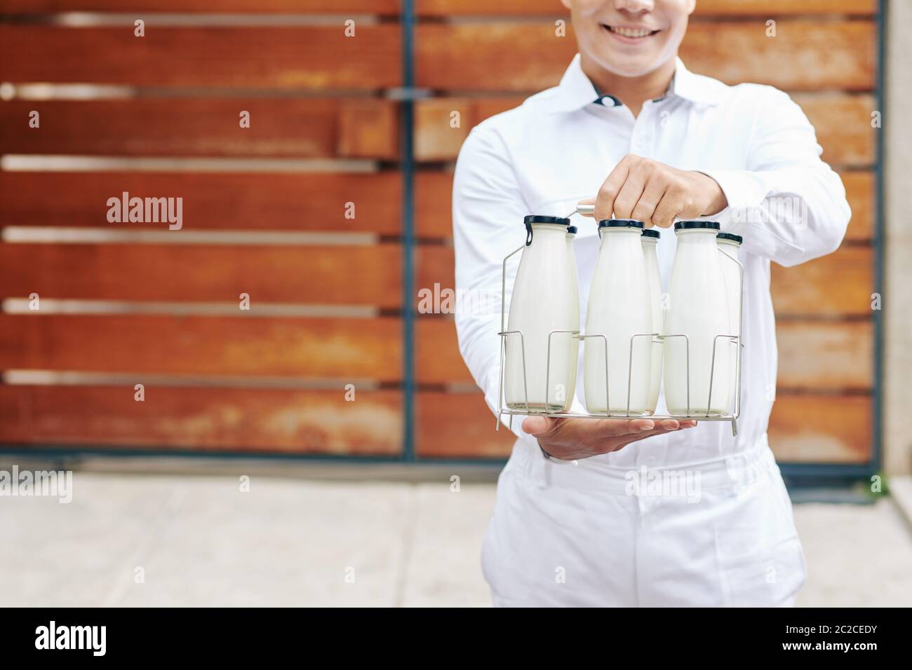 Young milkman in perfect white uniform standing against wooden fence holding glass bottles with milk, head cropped shot, copy space Stock Photo