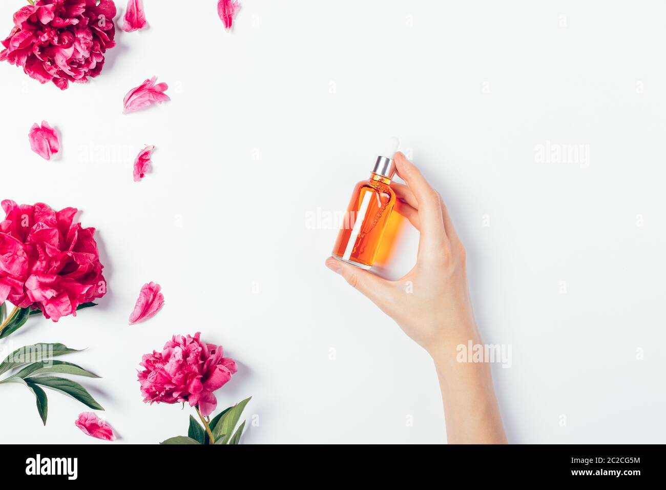 Female hand holding bottle of cosmetic serum next to fresh pink flowers on white background, flat lay. Stock Photo