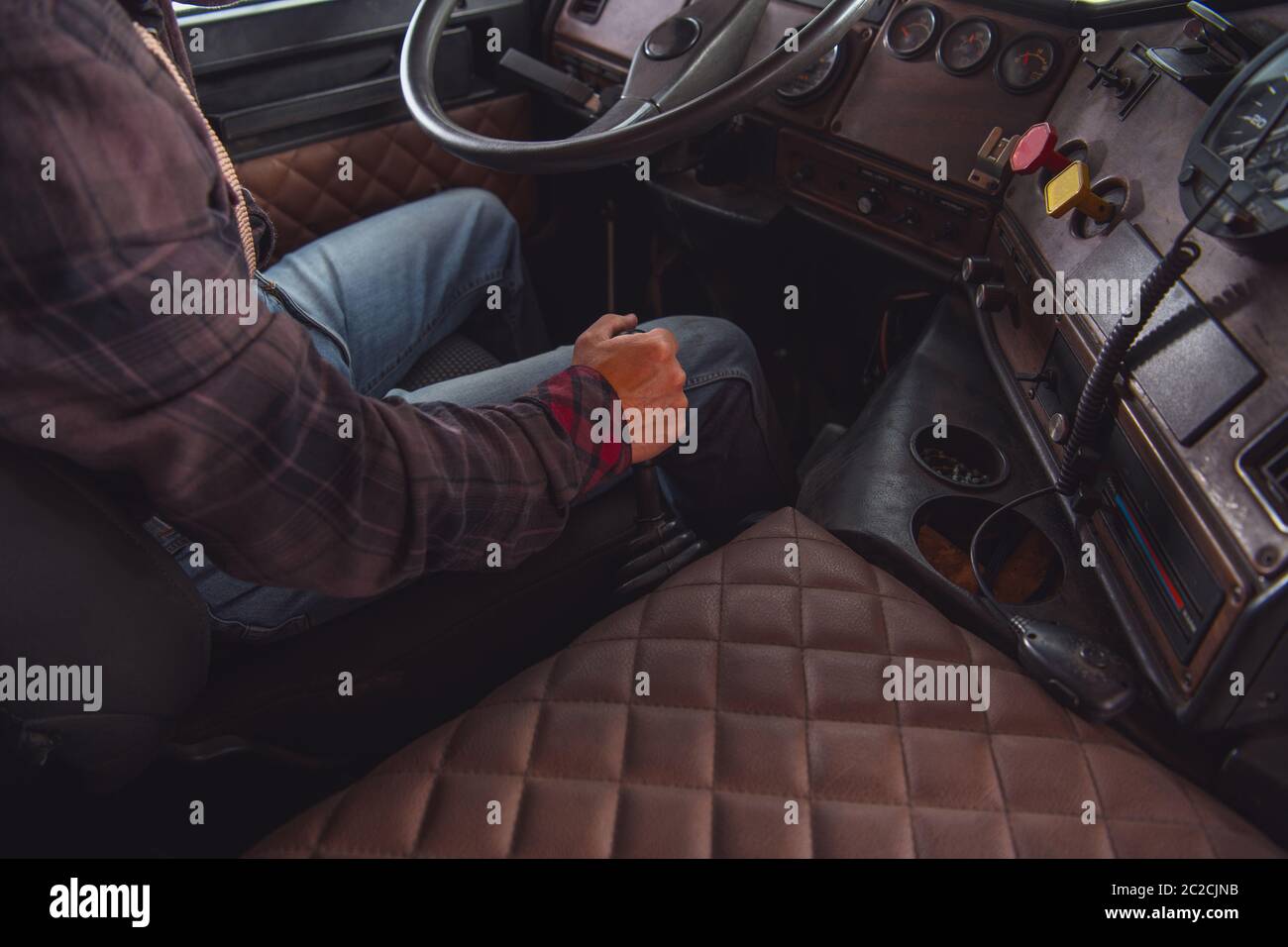 Caucasian Semi Truck Driver Behind the Steering Wheel. Manual Transmission in Aged Vintage Truck. Transportation Industry. Stock Photo