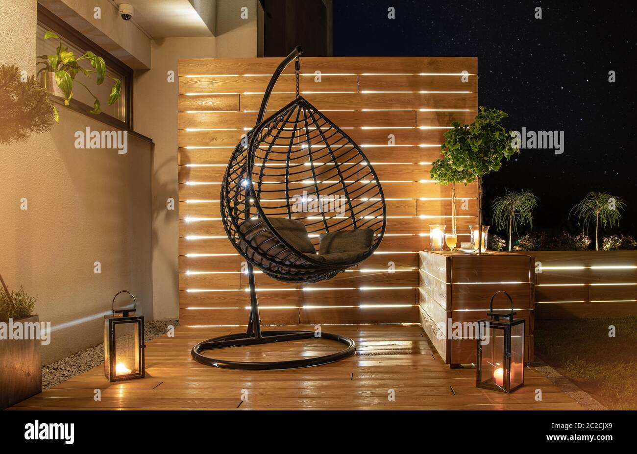 Garden Furniture Design. Starry Night Time in Stylish Backyard Garden with Wooden Porch with Wall and Planters Illuminated by Modern LED Lighting. Stock Photo