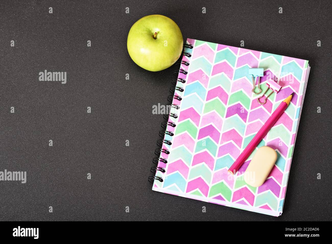 Top view of girly pink notebook with pencil, eraser, binder clips and apple on black background. Stock Photo