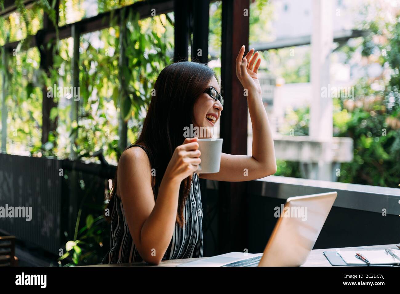 Smart young Asian woman smiling and waving hand while sitting at table with laptop and studying on terrace Stock Photo