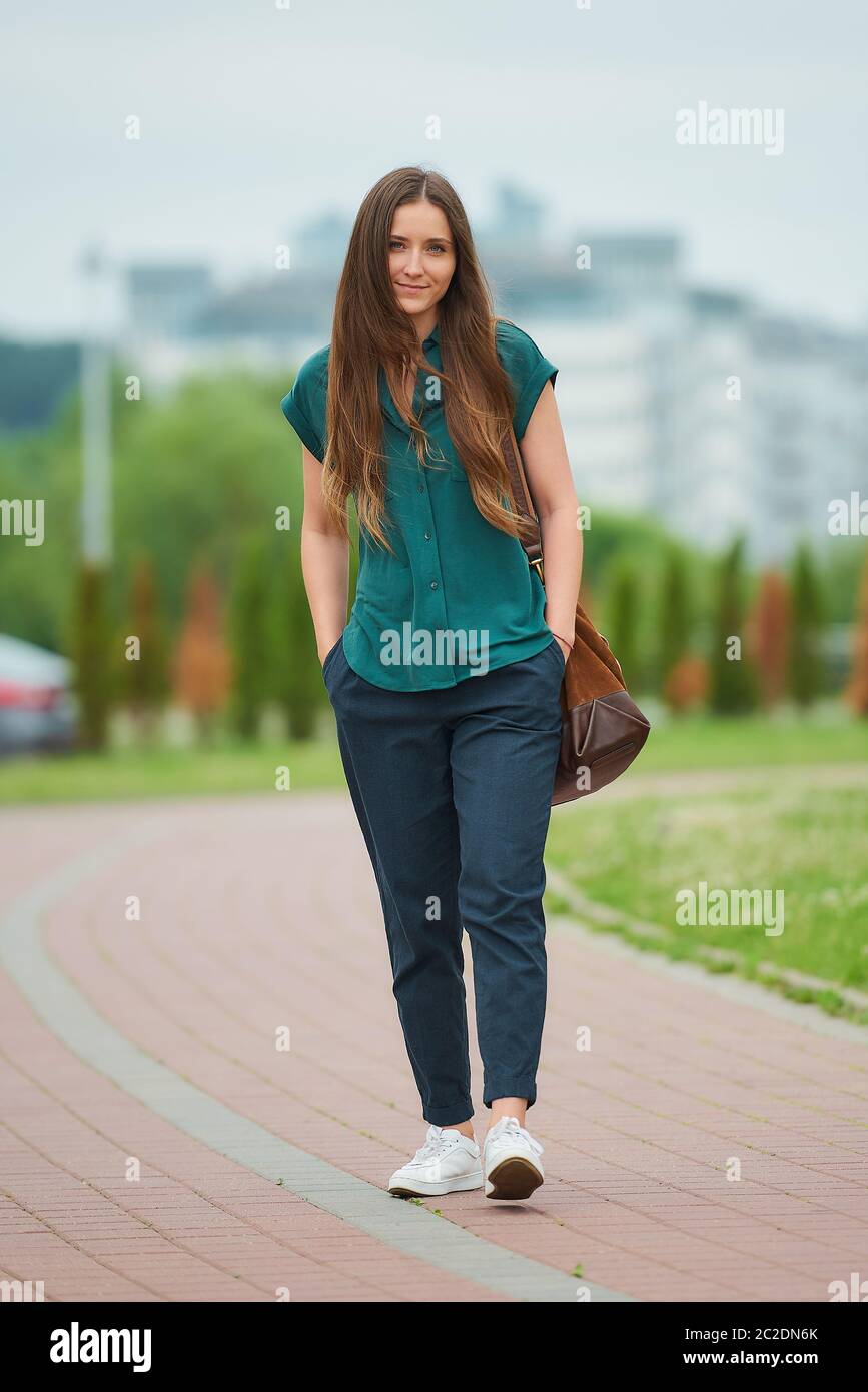 A young woman wears stylish casual clothes is walking thrusting hands into pockets of trousers in the park. A pretty girl wears office style clothes Stock Photo