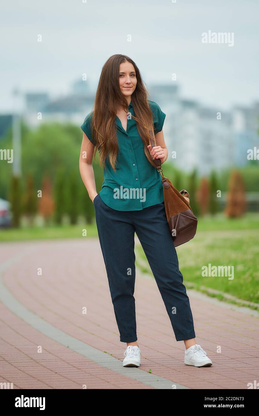 A young woman wears stylish casual clothes is walking thrusting a hand into a pocket of trousers in the park. A girl with long hair wears office style Stock Photo