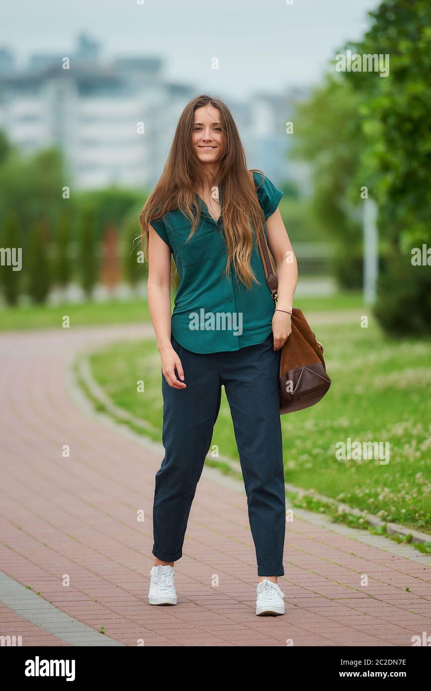 A young woman wears stylish casual clothes enjoying her walk in the park. A girl with long hair wears office style clothes keeping social distance Stock Photo