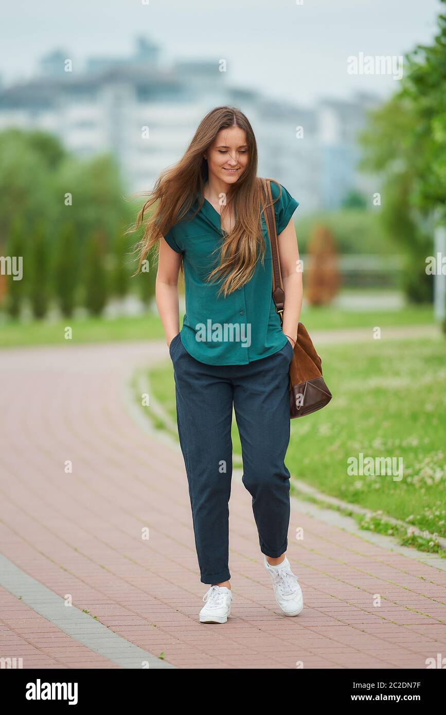 A woman wears stylish casual clothes enjoying her walk thrusting hands into pockets of trousers in the park. A pretty girl with long hair Stock Photo