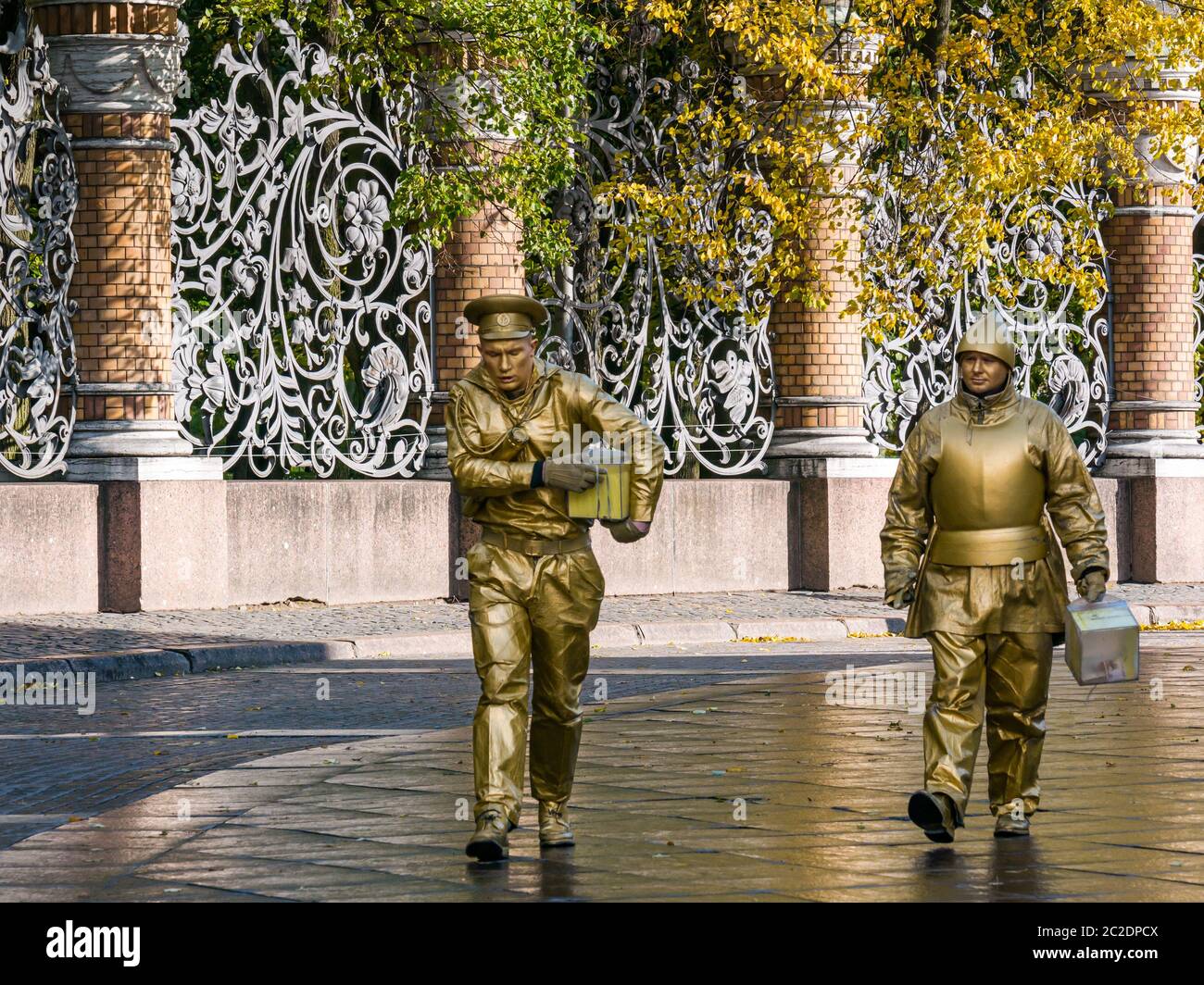 Men gold painted dressed as living statue tourist attractions walking past ornate metalwork fence of Mikhailovsky Garden, St Petersburg, Russia Stock Photo