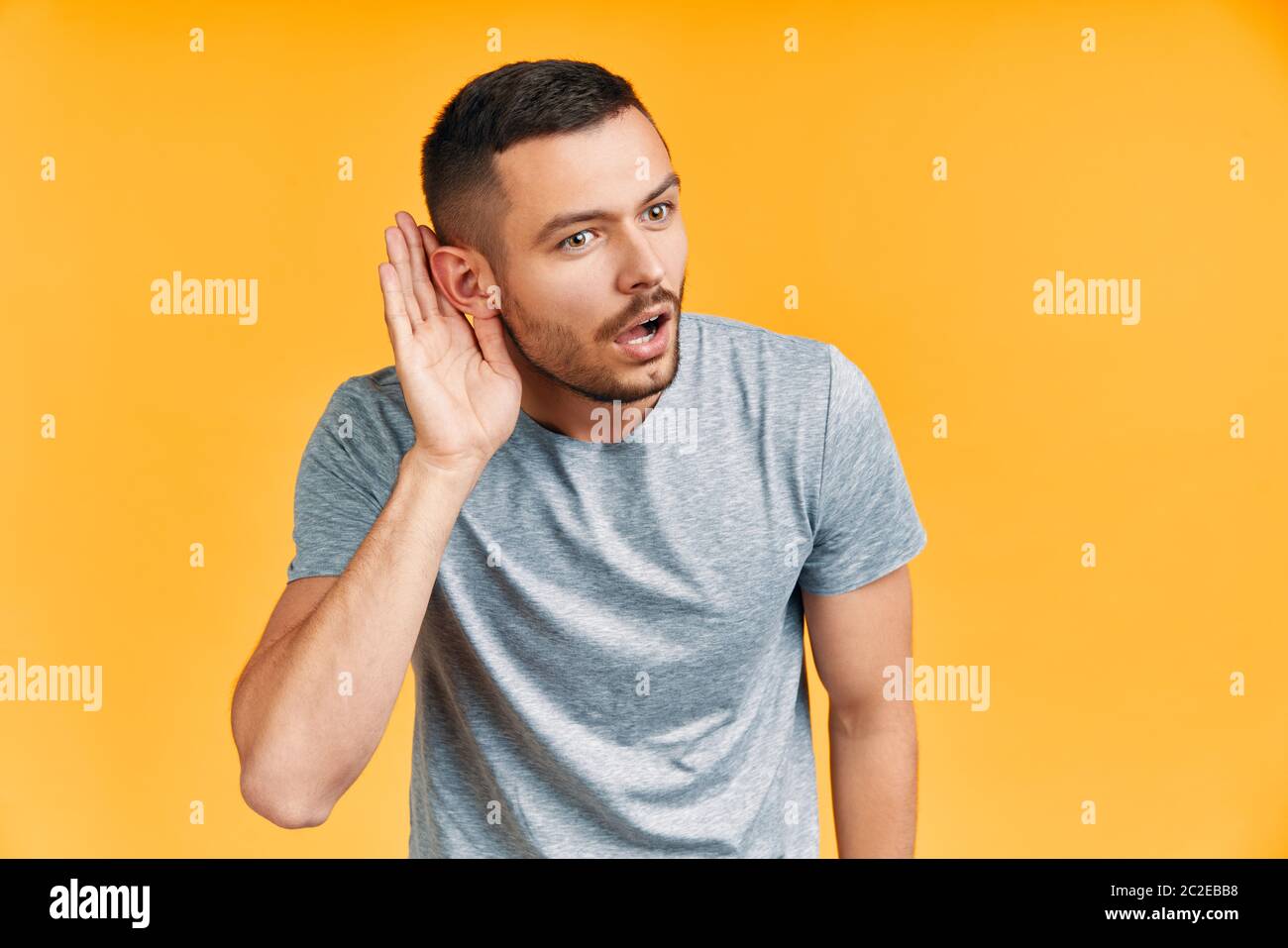 Young surprised man listening something carefully and holds his hand near ear over yellow background Stock Photo