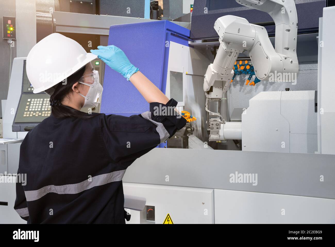 Female engineer wearing protective mask and glove to protect against Covid-19 and looking at robot CNC machine in smart factor Stock Photo