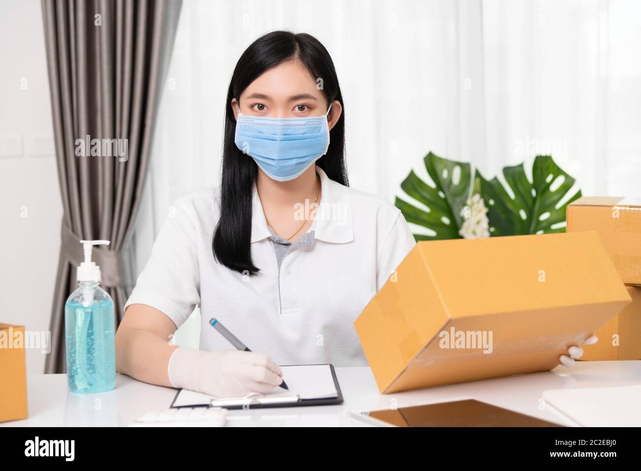 Young Asian small business owner wearing face mask protective to carrying product boxes at home office, startup SME entrepreneur or freelance woman wo Stock Photo