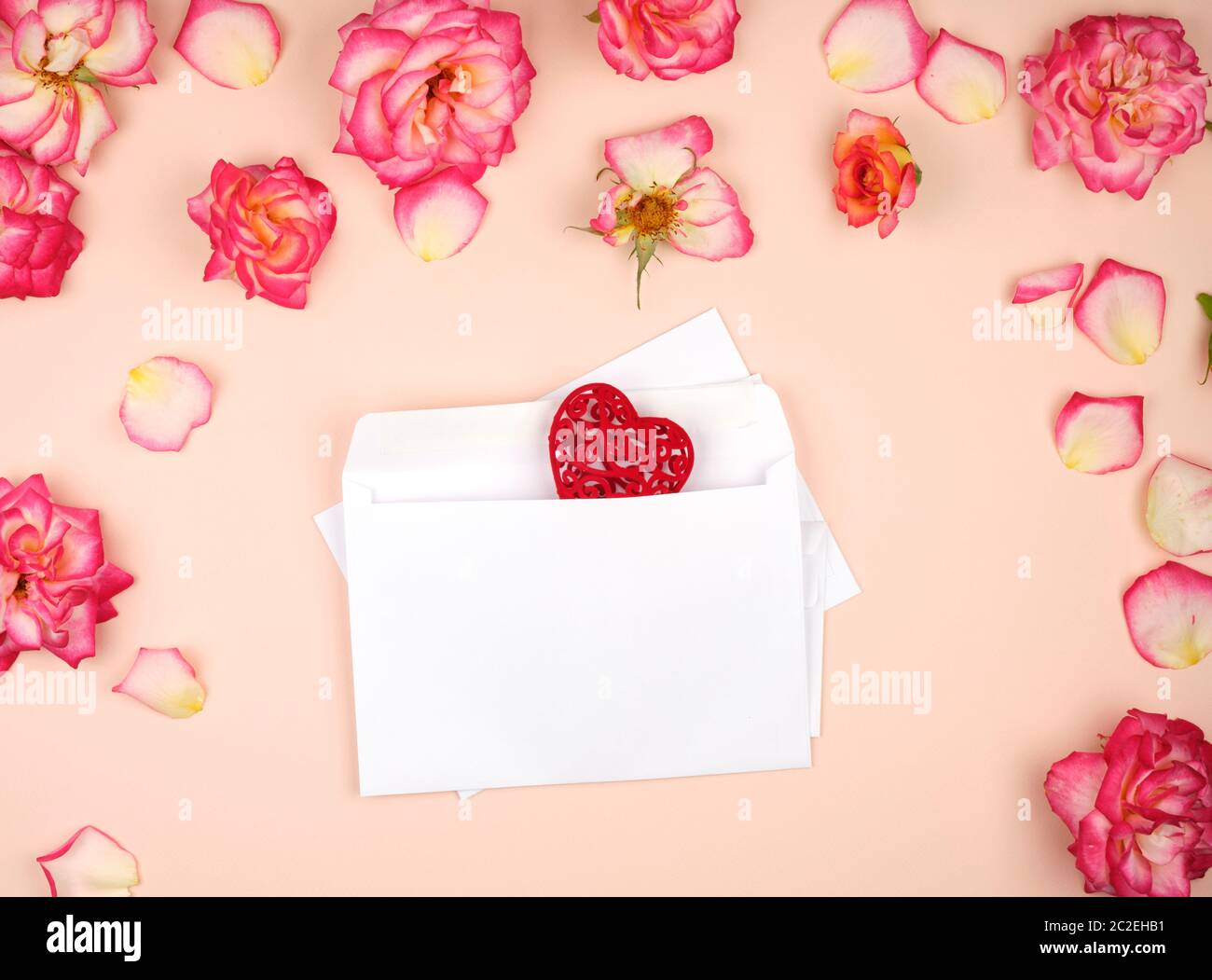 white paper envelopes and two red carved hearts in the middle of rosebuds, top view, festive backdrop Stock Photo