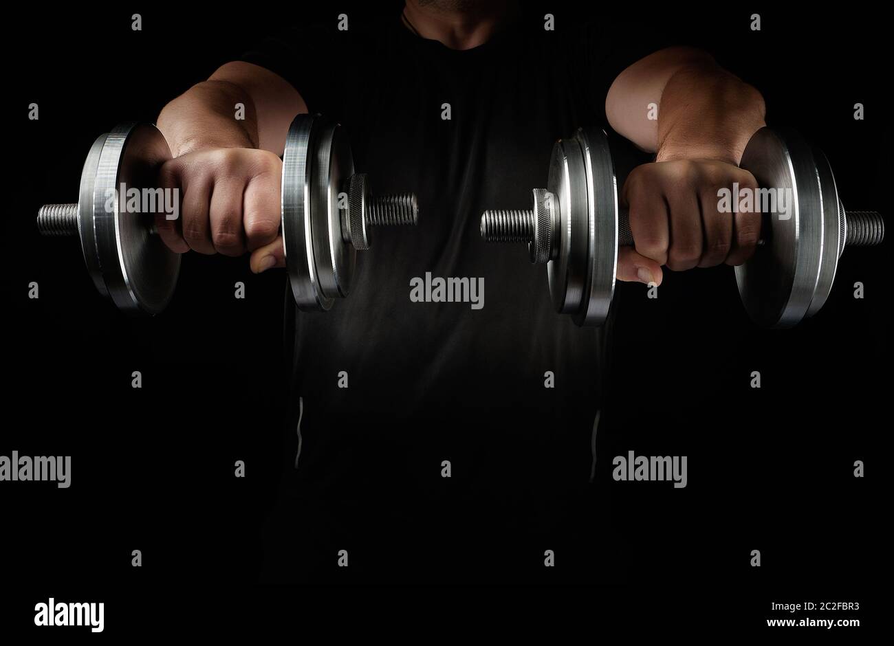 two steel typesetting dumbbells in male hands, arms extended forward and muscles tense, sports backdrop, low key Stock Photo