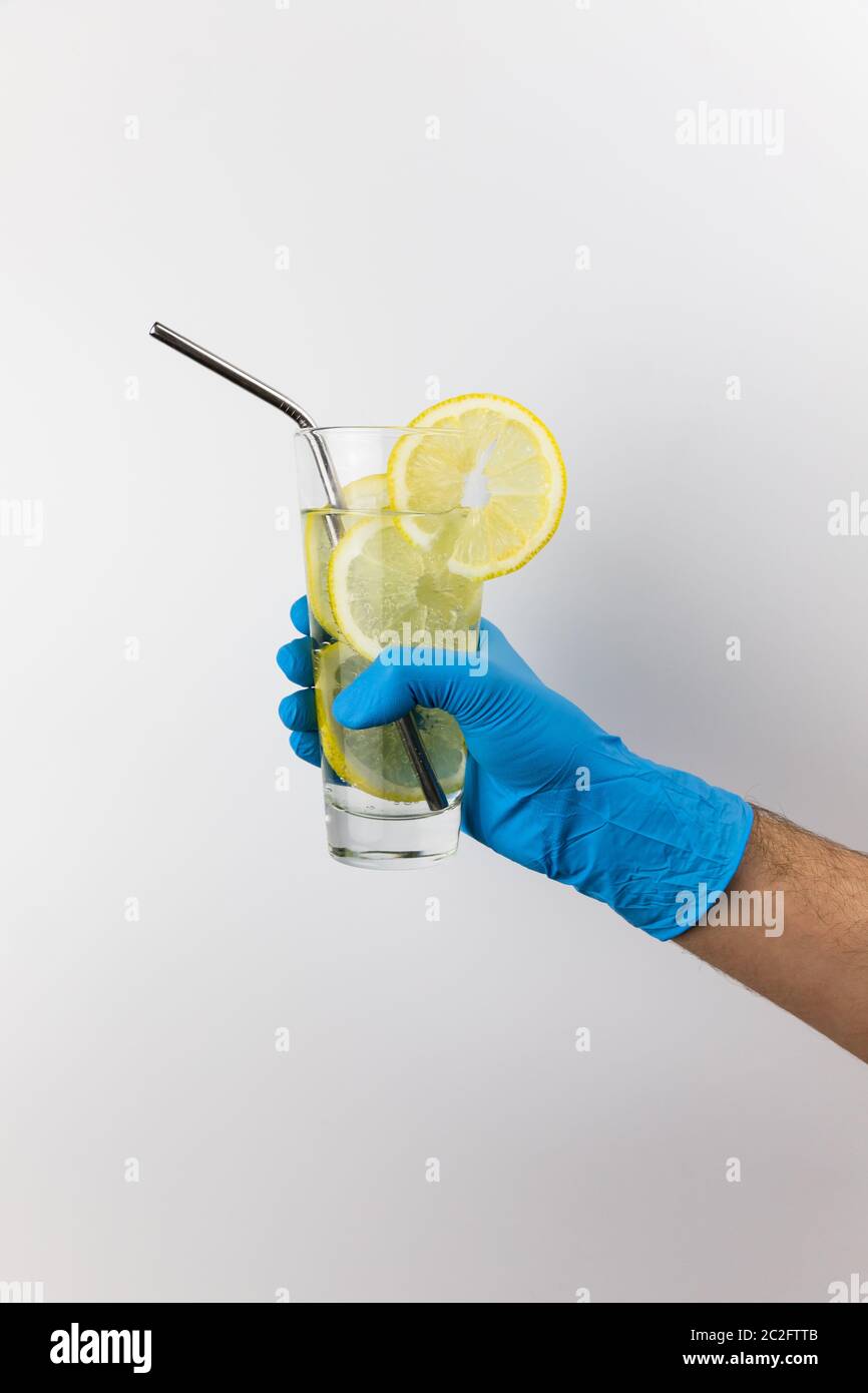 Man's hand holding a glass of cold sparkling water with lemon slices, wearing blue gloves on white background. Coronavirus pandemic prevention Stock Photo
