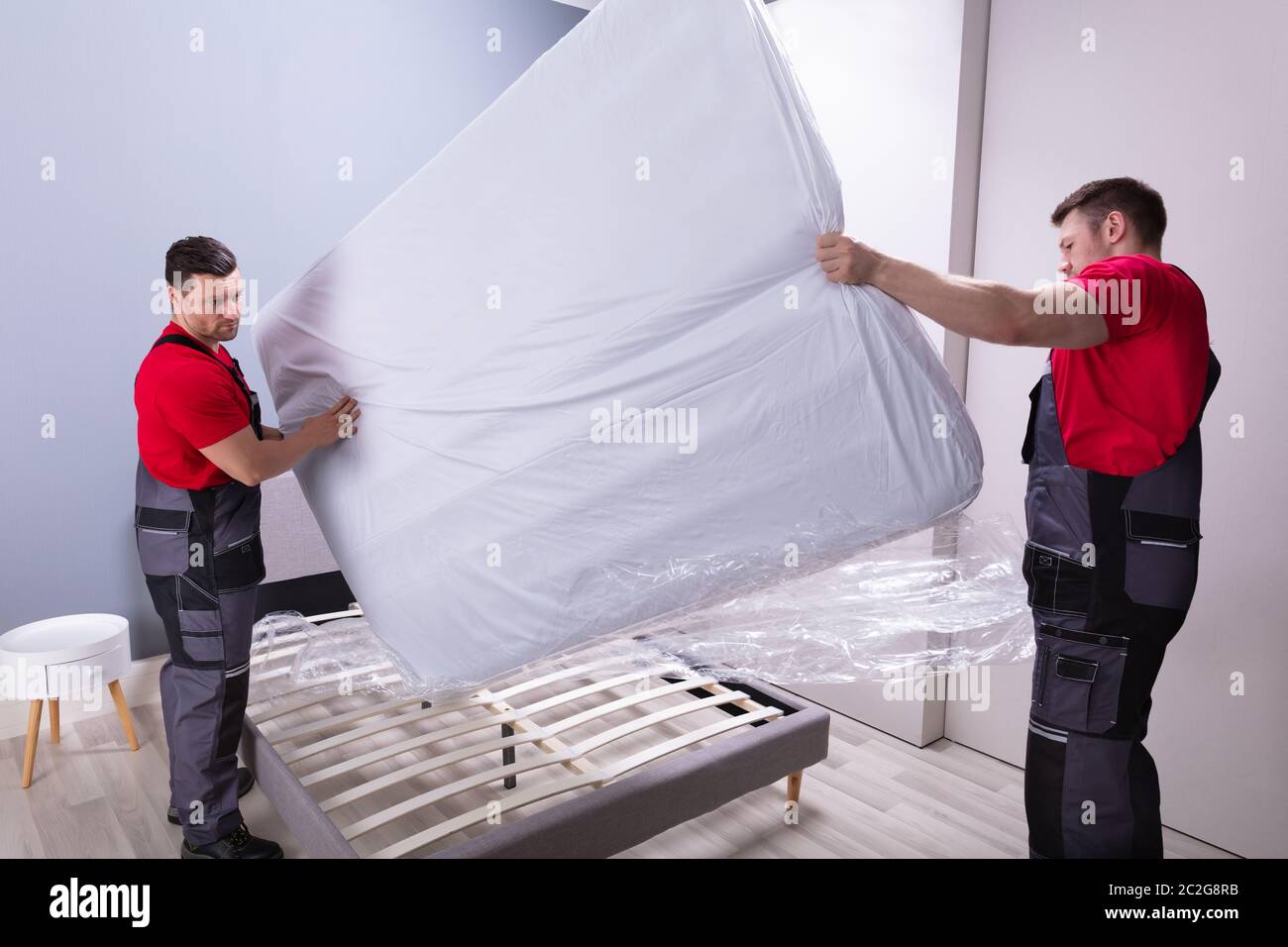 Two Male Professional Movers In Uniform Placing The Mattress Over The Bed In New House Stock