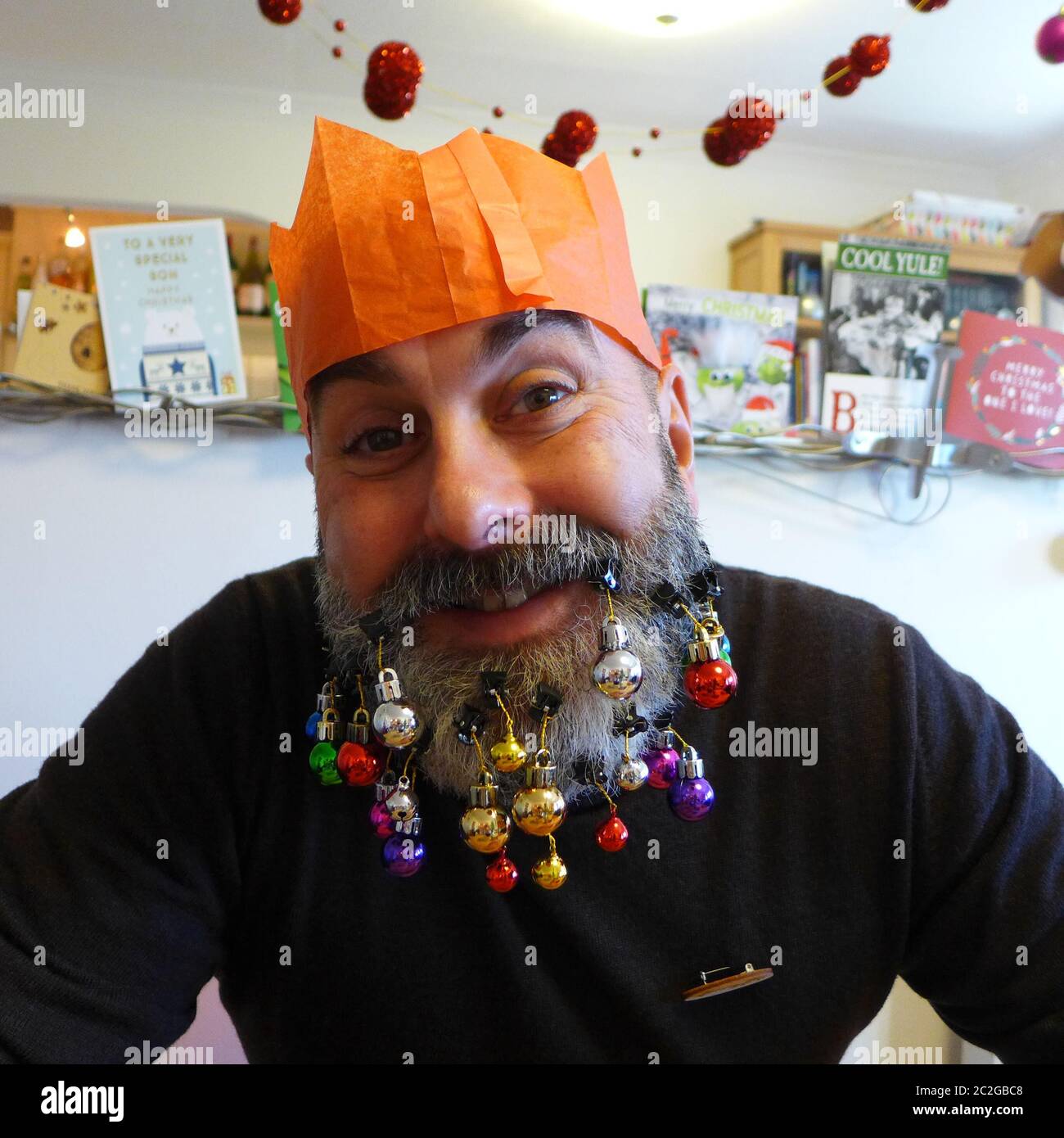 Christmas decorated beard with baubles, grey-haired man wearing paper hat from cracker looking bemused and happy Stock Photo