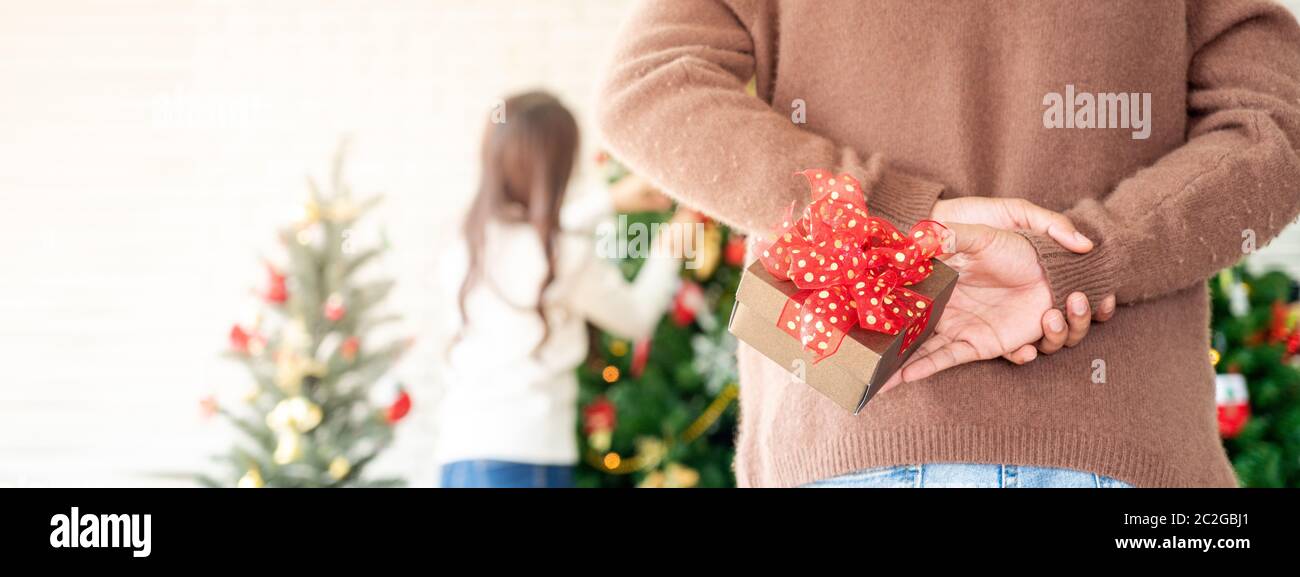 Man hold christmas gift behind his back for surprise his girlfriend while she decorating christmas tree in Chgristmas holiday season greeting. Panoram Stock Photo