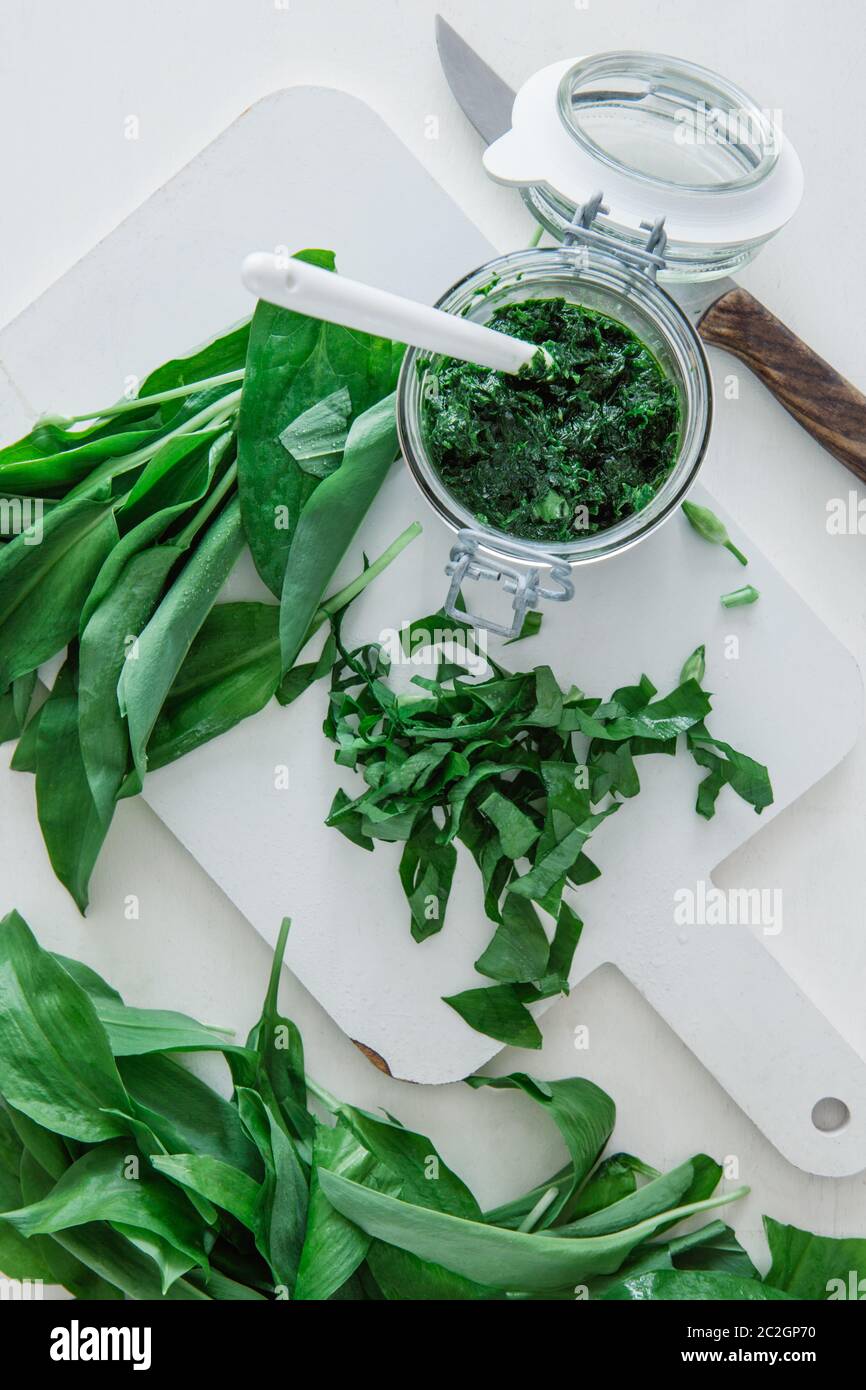 A glass of pesto and green leaves of wild garlic with kitchen utensils on a white table, top view Stock Photo
