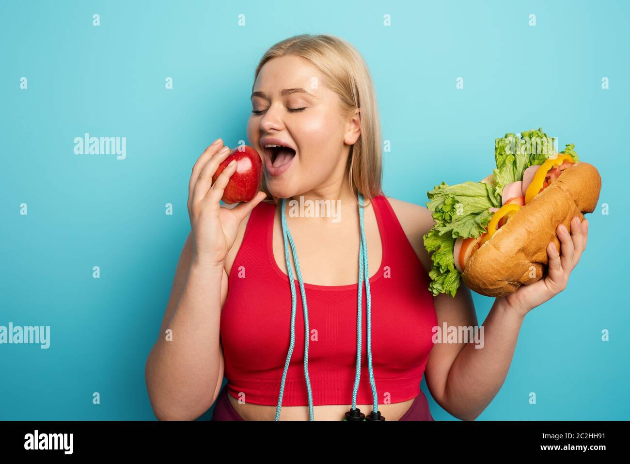 Fat girl prefers to eat an apple instead of a big sandwich. cyan background Stock Photo