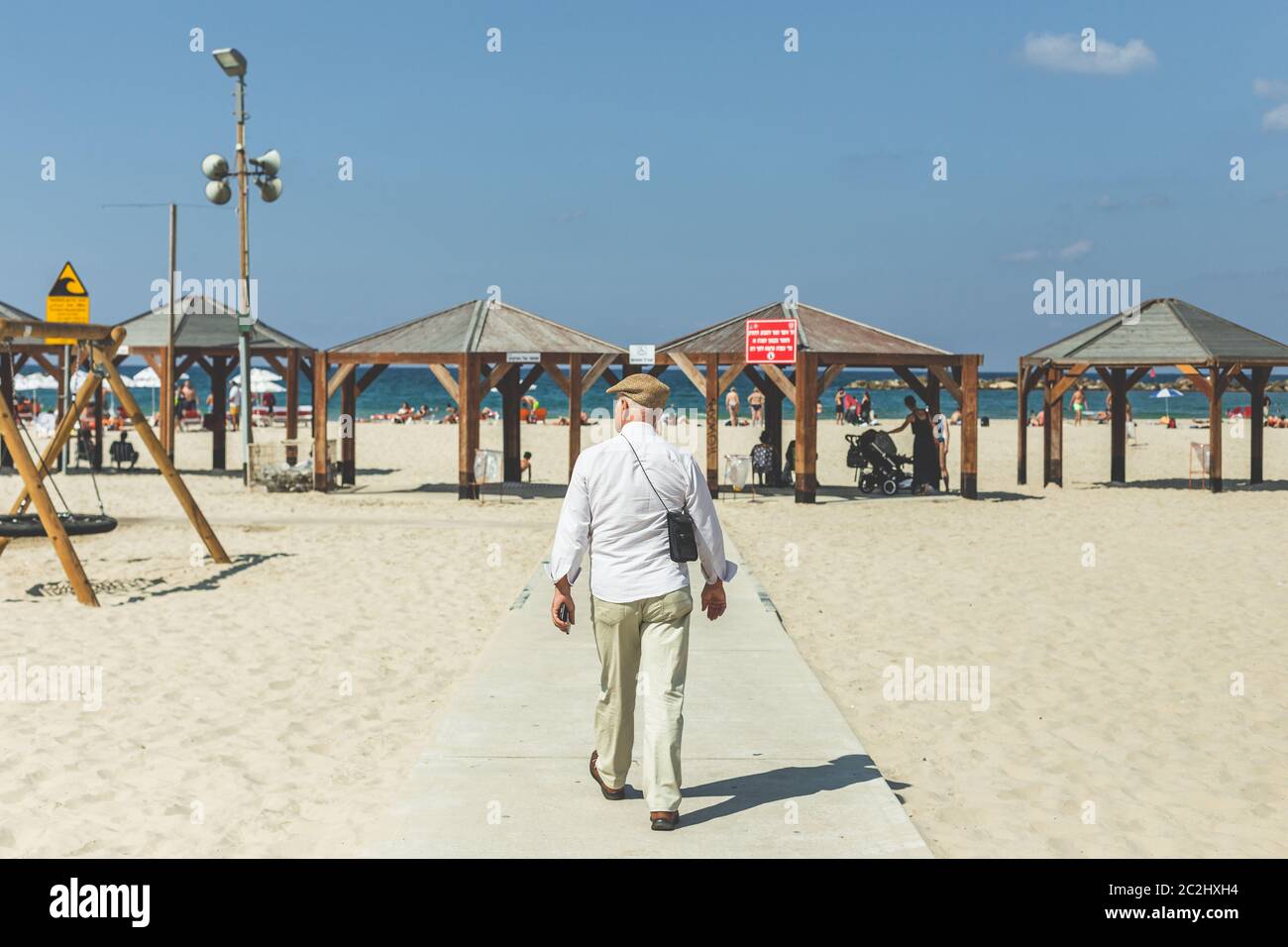 An older man in a flat cap, white shirt and trousers walking on a beach in Tel Aviv, Israel Stock Photo