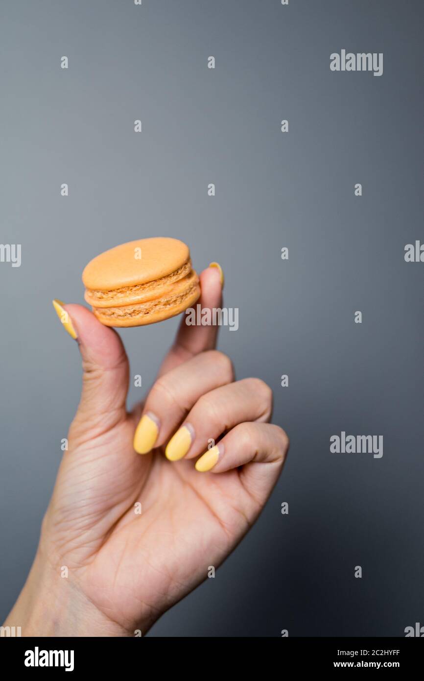 Close up of a woman's hand holding up an orange french macaron pastry on a dark background. Stock Photo