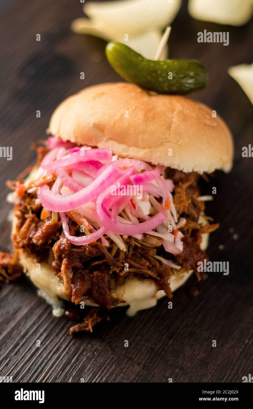 Slow cooked pulled pork with BBQ sauce, pickled onions, coleslaw and cheese served in a hamburger bun with dill pickle and chips as sides. It's messy. Stock Photo