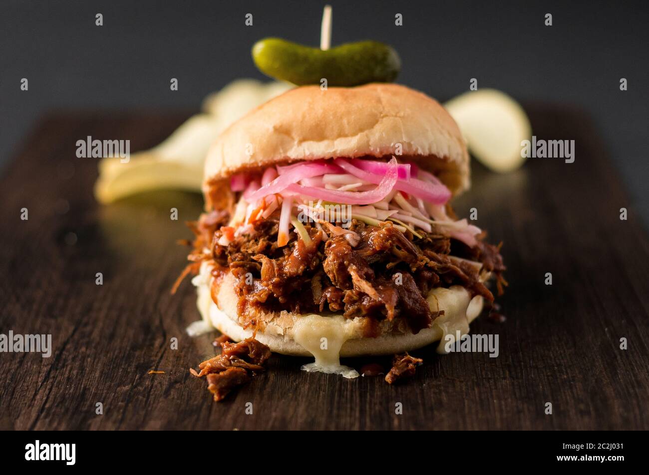 Slow cooked pulled pork with BBQ sauce, pickled onions, coleslaw and cheese served in a hamburger bun with dill pickle and chips as sides. It's messy. Stock Photo