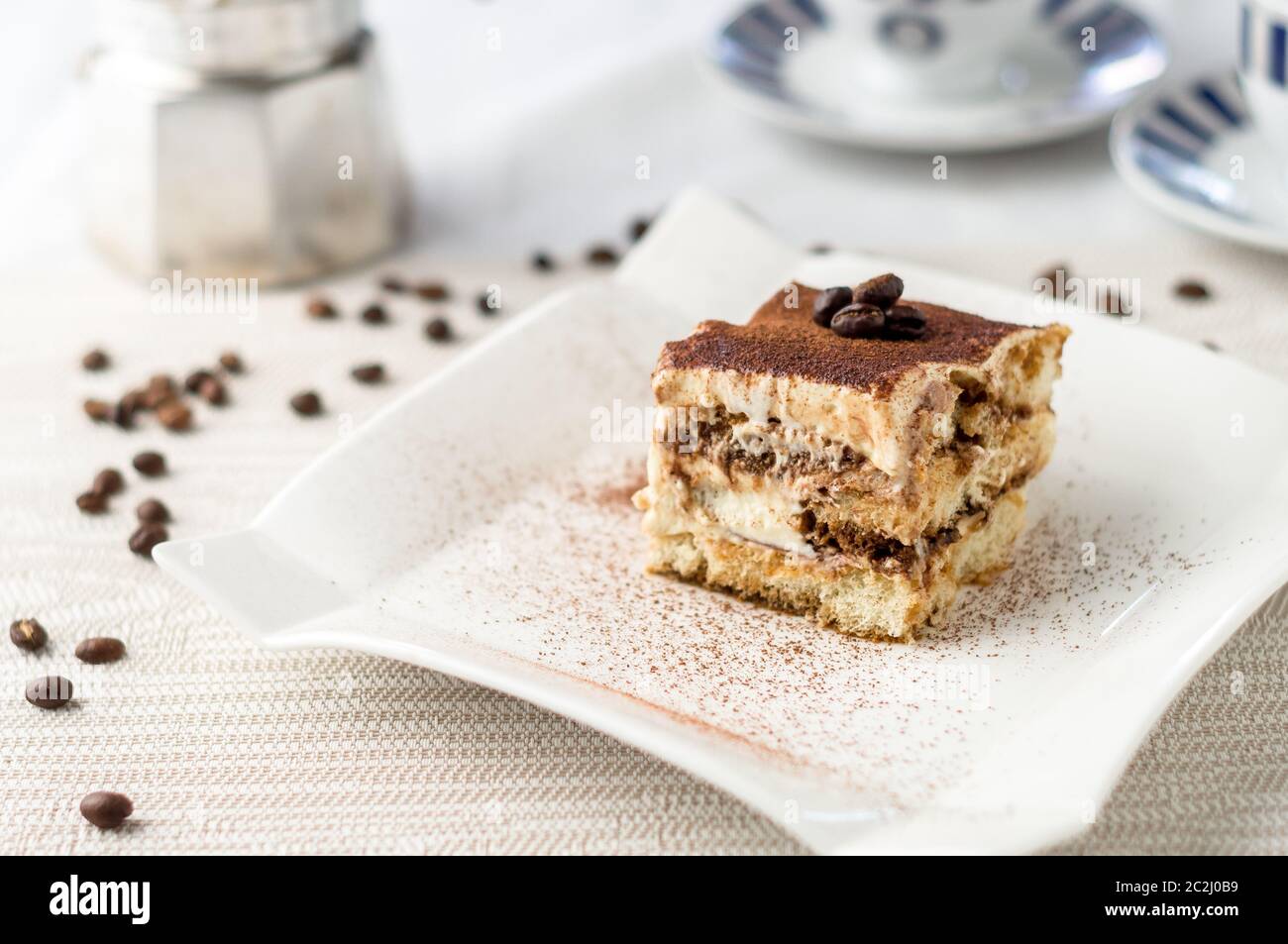 Delicious homemade tiramisu cake slice on a white plate. A classic Italian dessert. In the background espresso coffee cups and spilled coffee beans. Stock Photo