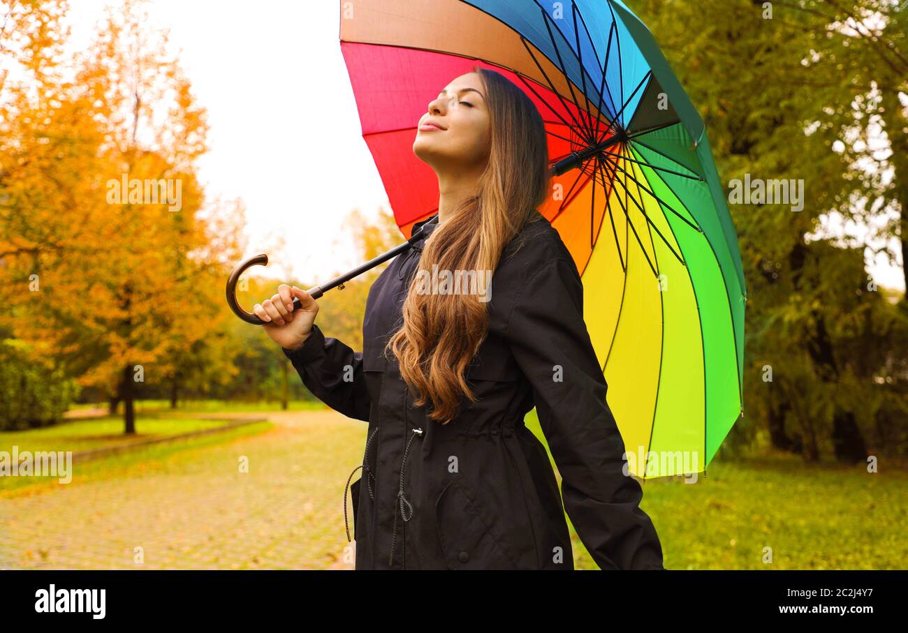 Portrait of a happy woman wearing raincoat under an umbrella breathing in city park on rainy day. Stock Photo