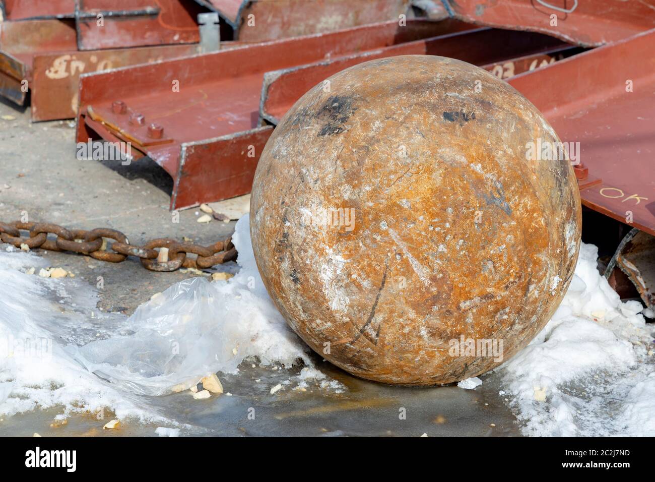 A rusty wrecking ball lying on the ground. A small amount of snow is around it, and steel beams are in the background. Stock Photo