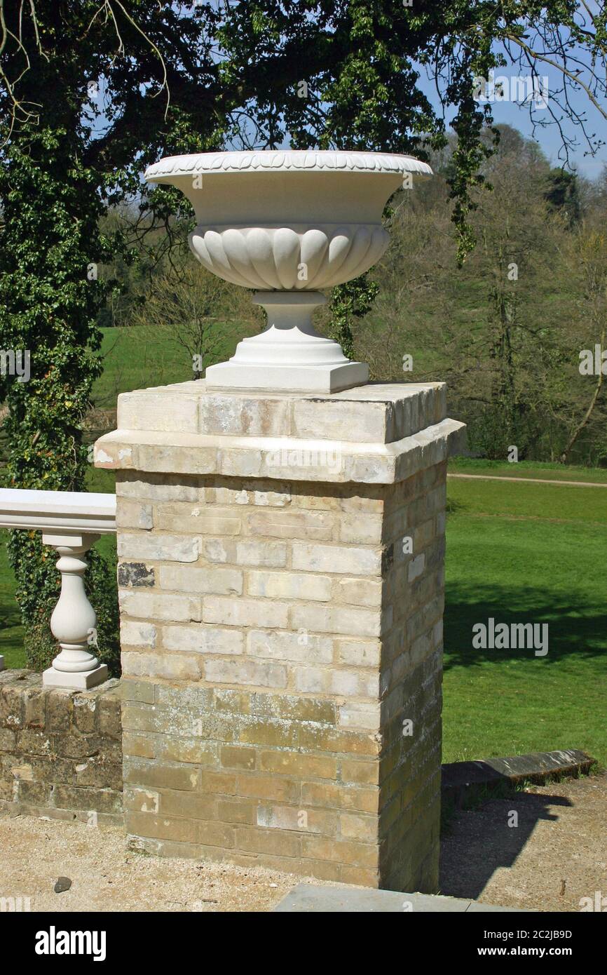 Ornamental jardiniere on a brick pillar attached to an old wall and balustrade constructed of light grey coloured bricks. Lawns, trees and blue sky in Stock Photo