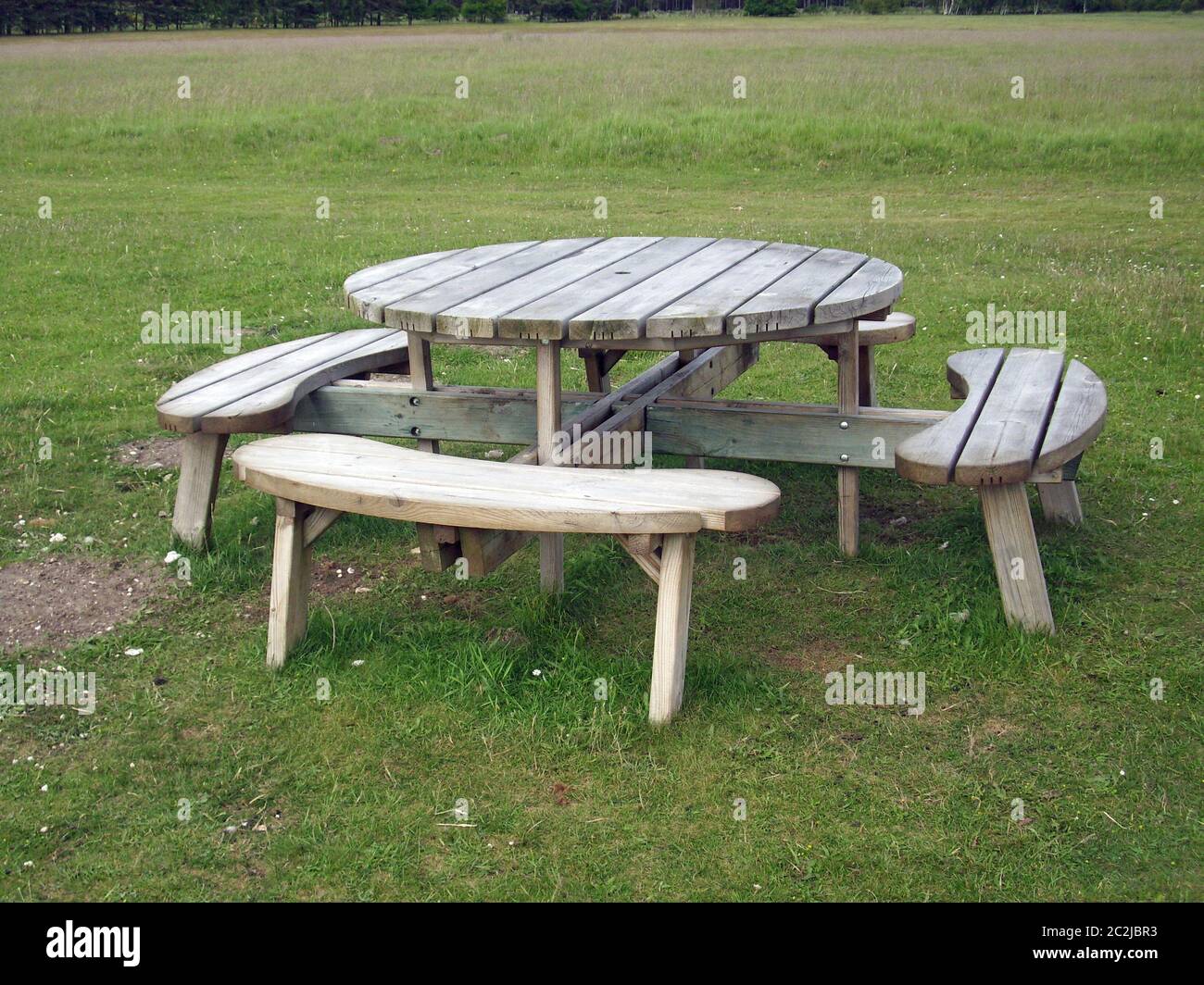 Round wooden picnic table with rounded seats on a grass field with trees  just visible in the background Stock Photo - Alamy