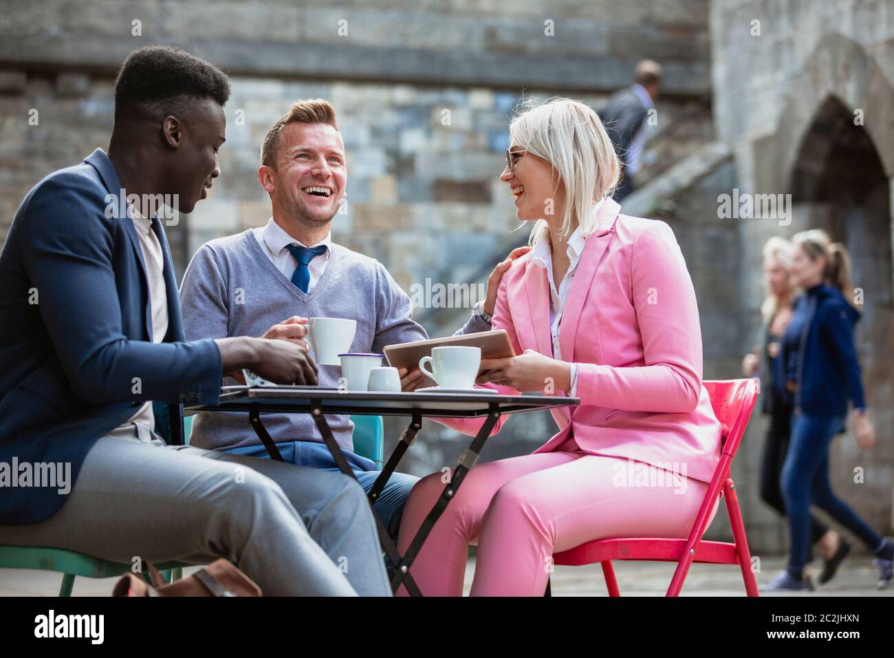 Two businessmen and a businesswoman sitting at an outdoor table in a city having a hot drink while laughing at something on a digital tablet. One man Stock Photo