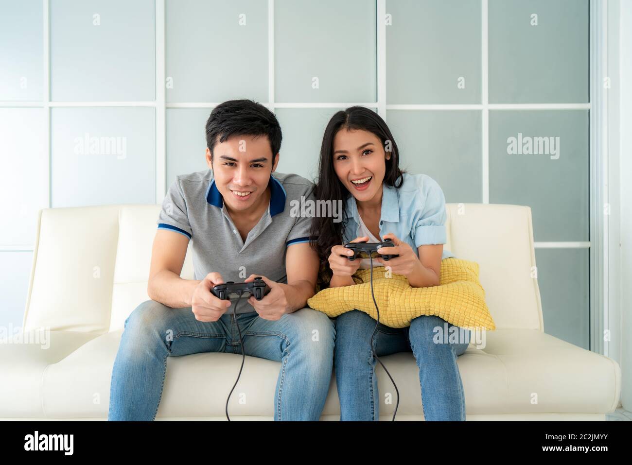 Asian couple man ans woman to playing video games with joysticks while sitting in sofa in living room at home, concept of family values and the modern Stock Photo